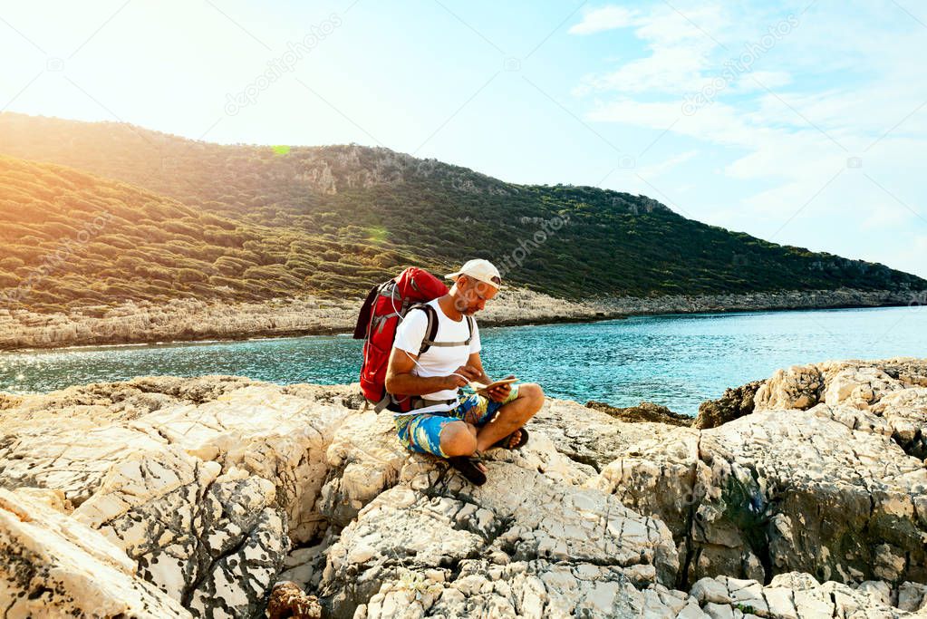 Traveler with a portable solar battery attached to his backpack is sitting on a rocky seashore and using tablet pc - adventure travel along Lycian way, Turkey. Backlight.