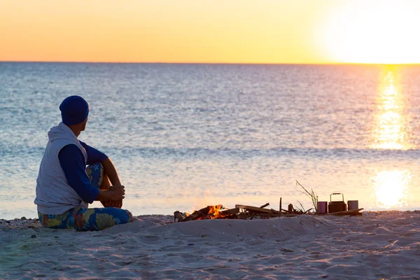 海岸で焚き火の隣に旅行者の座っている瞑想し 海の向こうの夕日を賞賛します 海辺の素晴らしい旅 背面図 — ストック写真