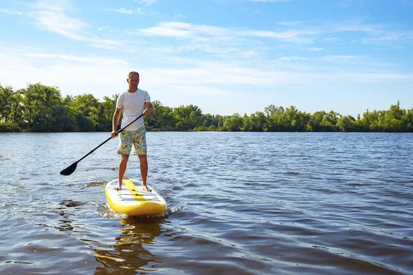 Happy man is paddling on a SUP board on a large river at sunny day. Stand up paddle boarding - awesome active recreation in nature.