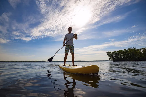 Happy man is paddling on a SUP board on a large river at sunny day. Stand up paddle boarding - awesome active recreation in nature.  Wide angle.