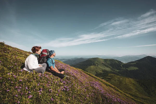 Travelers, friends relax on the mountain trail among flowering pink rhododendrons. Epic travel in the mountains. Back view, wide angle, vintage image. Cosmic atmosphere.