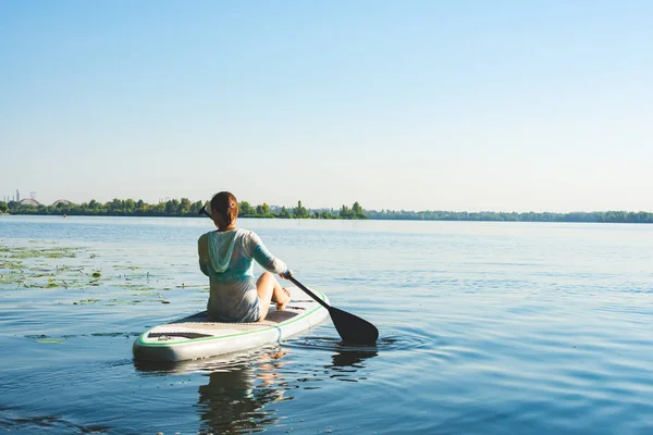 Happy woman relaxes on SUP board and enjoy life. Stand up paddle boarding - awesome active outdoor recreation. Back view.