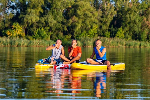 Joyful friends, a SUP surfers relax, eat apples and having fun during rest in nature. Stand up paddle boarding - awesome active outdoor recreation.