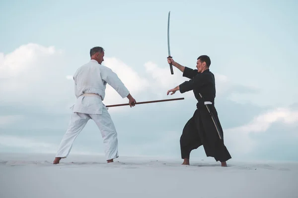 Two men, in Japanese clothes, are practicing martial arts with a traditional Japanese weapon - a katana and jo, in the desert at sunset.