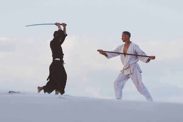 Two men in Japanese clothes are practicing martial arts with a traditional Japanese weapon - a katana and jo  in the desert at sunset.