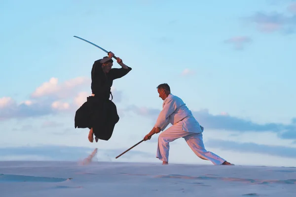 Japon kıyafetli konsantre erkeklerde bir silahla geleneksel Japon - bir katana ve jo, gün batımında çölde dövüş sanatları uygulamak.