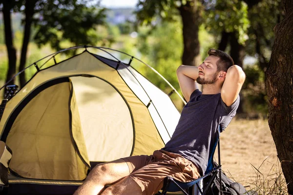 Een man rust in de buurt van een tent op een klapstoel in het bos. — Stockfoto