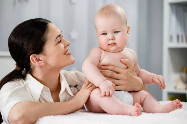 Mãe suporta um bebê nu que se senta em uma fralda em um quarto brilhante — Fotografia de Stock