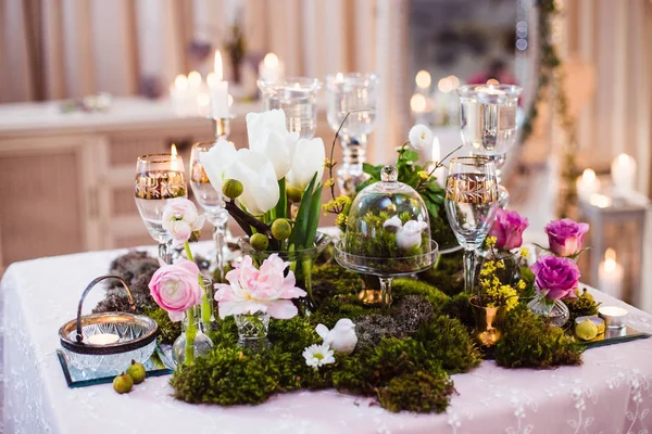 A table for two. Romantic atmosphere in subdued light, lighted candles and rustic decoration (forest moss, flowers, glasses and vases)