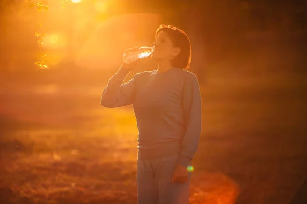 Elderly woman Drinking water from a bottle at sunset — Stock Photo, Image