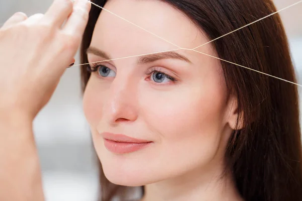 Cosmetologist plucks client eyebrows by thread. Close up