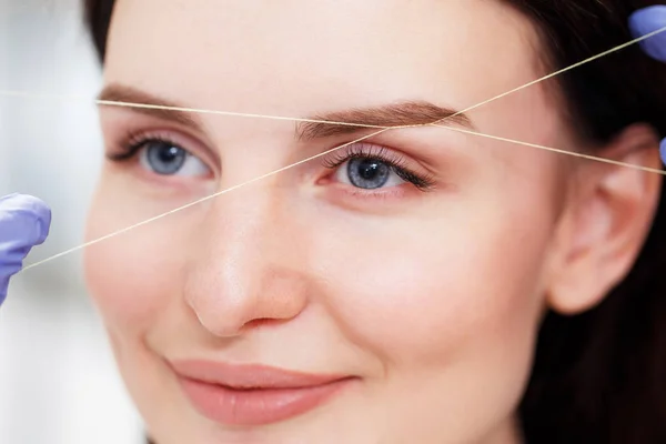 Cosmetologist plucks client eyebrows by thread. Close up