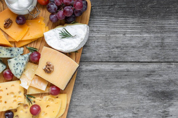 Assorted cheeses on wooden board plate served with walnuts, grapes and rosemary on rustic wood background, top view with copy space