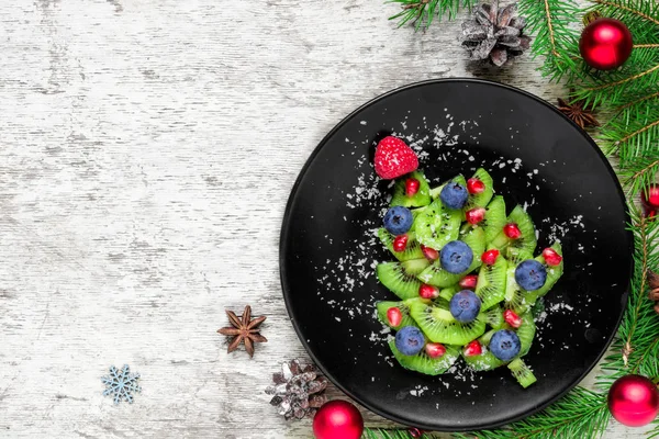 kiwi christmas tree with berries and coconut with fir tree branches and decorations over white wooden table. funny food idea for kids. Christmas and New Year food background. top view with copy space