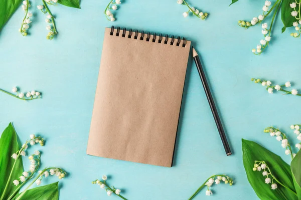 Blanco papieren notitieboekje in frame gemaakt van Lily of the Valley met een potlood op blauwe houten tafel. platte lag. Top View — Stockfoto