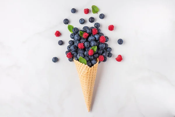 wild berries blueberry and raspberry with mint in waffle ice cream cone on white marble background. summer food concept