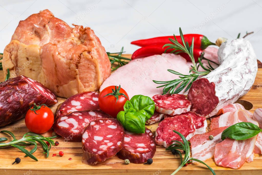 Cutting board with french salami, smoked sausage, bacon, tomatoes and herbs on white marble background