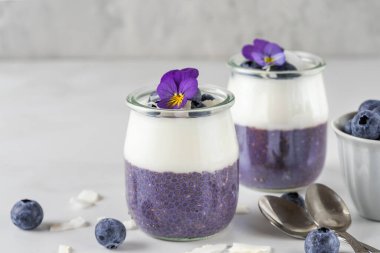 Taze böğürtlenli yaban mersinli chia pudingi, hindistan cevizli yoğurt ve beyaz masada yenebilir çiçekli sağlıklı vejetaryen kahvaltısı.