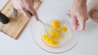 Beyaz mutfak masasındaki cam kasede çiğ yumurta çırpan bir adam. Kahvaltı hazırlıkları sırasında biber öğütücü. Omlet ya da çırpılmış yumurta. Sağlıklı yiyecekler. Üst görünüm.