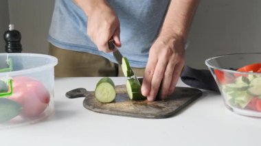 Man slicing fresh cucumber on cutting board with vegetables and glass bowl on kitchen table during salad preparation. Cooking healthy vegan food. Plant based eating, home cooking.