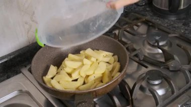 Seasoned potato slices being added to frying pan with oil on gas stove during meal preparation. Cooking french fries process on stovetop at home. Everyday culinary preparation.