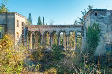 Abandoned colonnade in Tskaltubo surrounded by overgrown vegetation and decaying buildings under blue sky, Georgia. Haunted house. Post apocalyptic, creepy and spooky, harmony of ruin and nature.