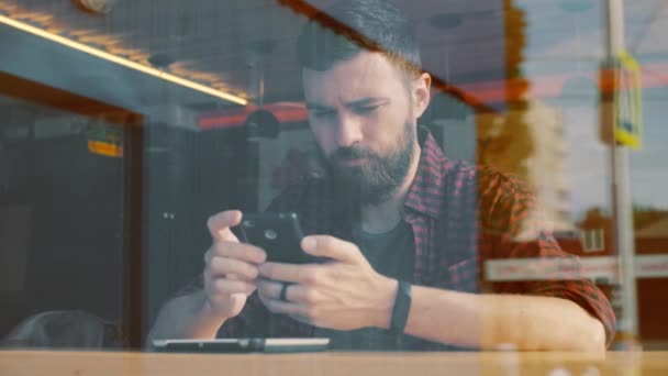 Hombre usando la aplicación en el teléfono inteligente en la cafetería. Disparo a través de la ventana — Vídeos de Stock