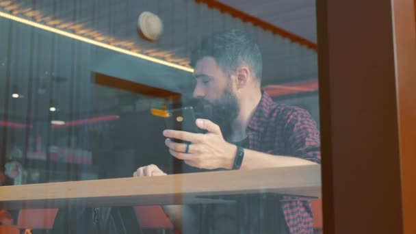 Βολή του ένας άνθρωπος που κάθεται στο καφενείο με ένα smartphone. Βολή μέσα από καφετέρια Εμφάνιση παραθύρου. — Αρχείο Βίντεο