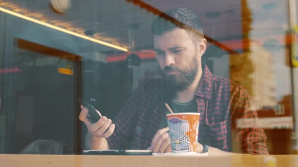 Bir kadeh bir akıllı telefon ile kafede oturan bir adam. Shot Cafe aracılığıyla penceresini göster. — Stok video