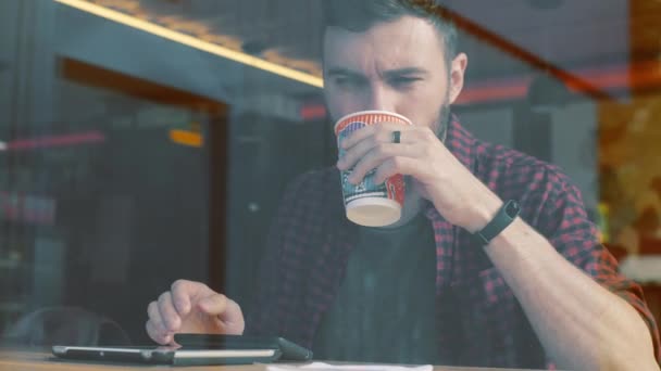 Tiro de um homem sentado no café com um smartphone. Shot através da janela de show de café . — Vídeo de Stock