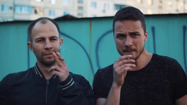 Двое мужчин курят сигареты в гетто и смотрят в камеру — стоковое видео