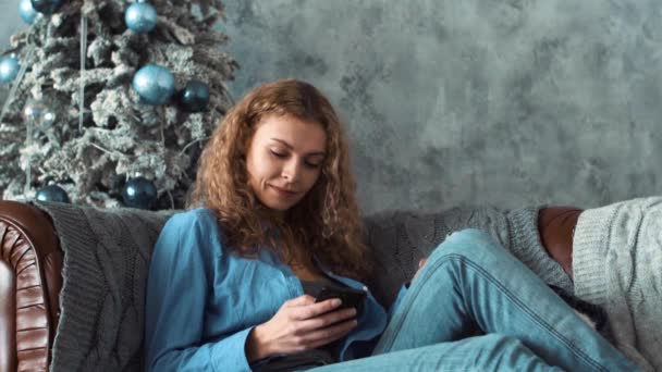 自宅のソファと彼女の携帯電話にテキスト メッセージの上に座って魅力的なブロンドの女の子 — ストック動画