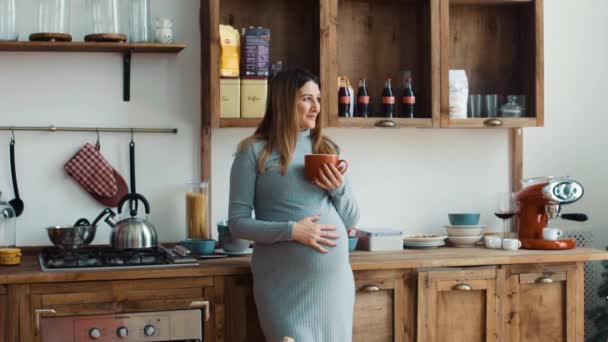 Mujer embarazada en la cocina está bebiendo de una taza roja — Vídeo de stock