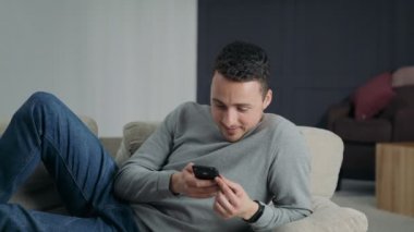 Mutlu genç adam kullanır Smartphone Lying bir kanepe evde iken