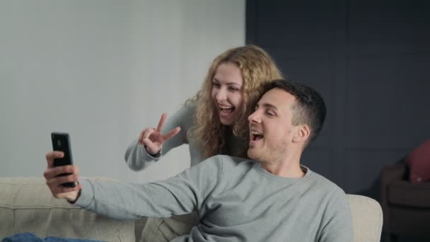 Пара, делающая селфи на камеру смартфона, сидящая и улыбающаяся дома — стоковое видео