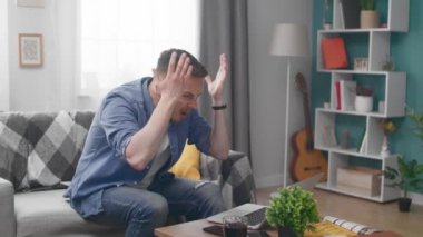 Young man sits on the couch and gets angry because of the bad news on the laptop
