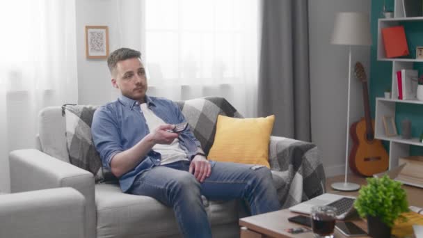 Young man sitting bored on the sofa, watching TV and changing channels — Stock Video