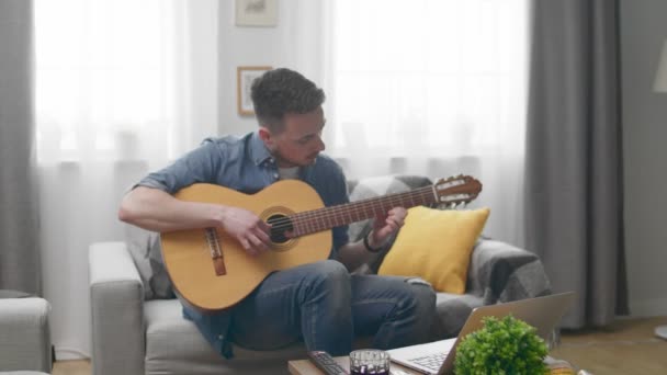 Bonito homem tocando uma guitarra acústica em casa no sofá — Vídeo de Stock