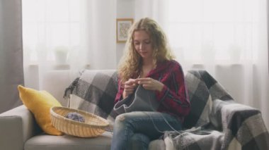Zoom in of young woman sitting on couch and knitting in home