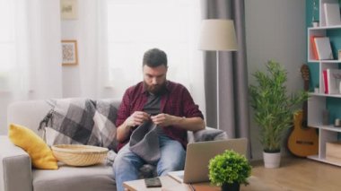 Kanepede oturan ve dizüstü bilgisayarda örgü öğretici izlerken genç adam Panning