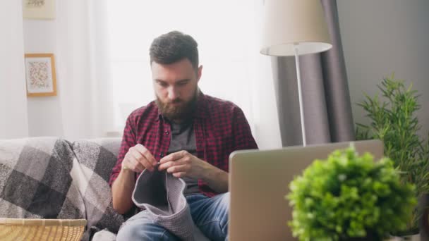 Handhållen av ung man sitter på soffan och tittar stickning tutorial på laptop — Stockvideo