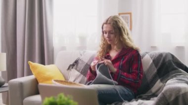 Zoom in of woman sitting on sofa and knitting crochet clothes in cozy home