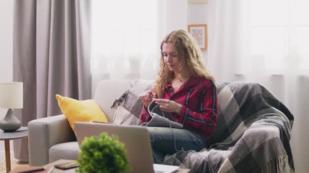 Средний снимок женщины, сидящей на диване и вязающей вязаную одежду в уютном доме — стоковое видео