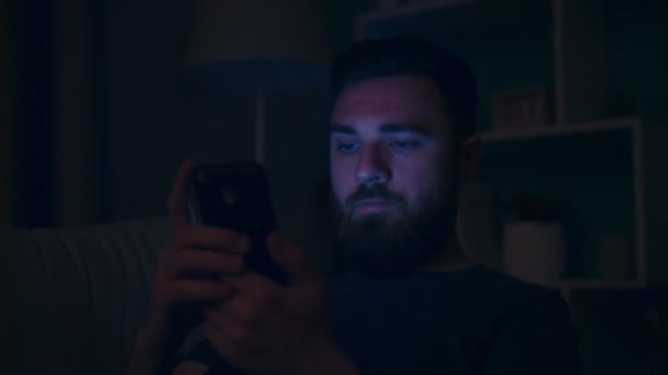 Вечером мужчина средних лет сидит на диване и пользуется смартфоном — стоковое видео