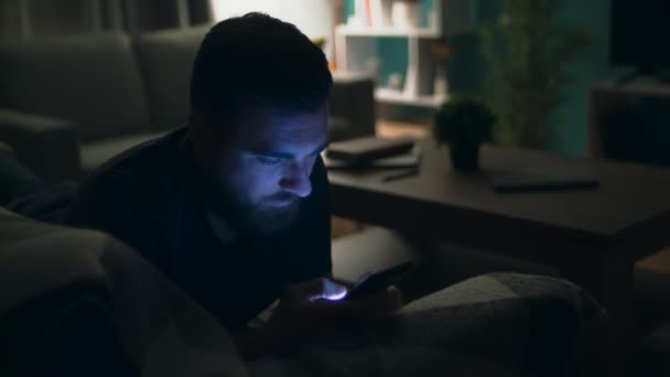 Joven hombre viendo la película en el teléfono inteligente acostado en el sofá en casa por la noche — Vídeos de Stock