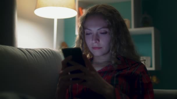 Hermosa mujer joven utiliza el teléfono inteligente en un sofá en la habitación acogedora por la noche — Vídeo de stock