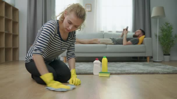 Müde Frau putzt Parkettboden und ihr Mann liegt mit Smartphone auf Sofa — Stockvideo
