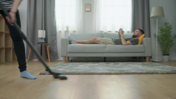 Уставшая жена пылесосит паркетный пол, а ее муж играет на смартфоне на диване — стоковое видео