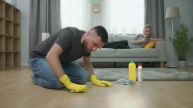 Erkek orta çekim parke zemin temizler, ve kadın akıllı telefon ile kanepe üzerinde yatıyor