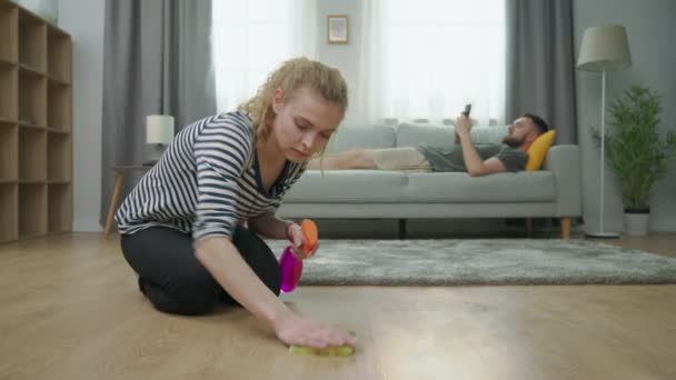 Tired wife cleans parquet floor and her husband lying on sofa with smartphone — Stock Video